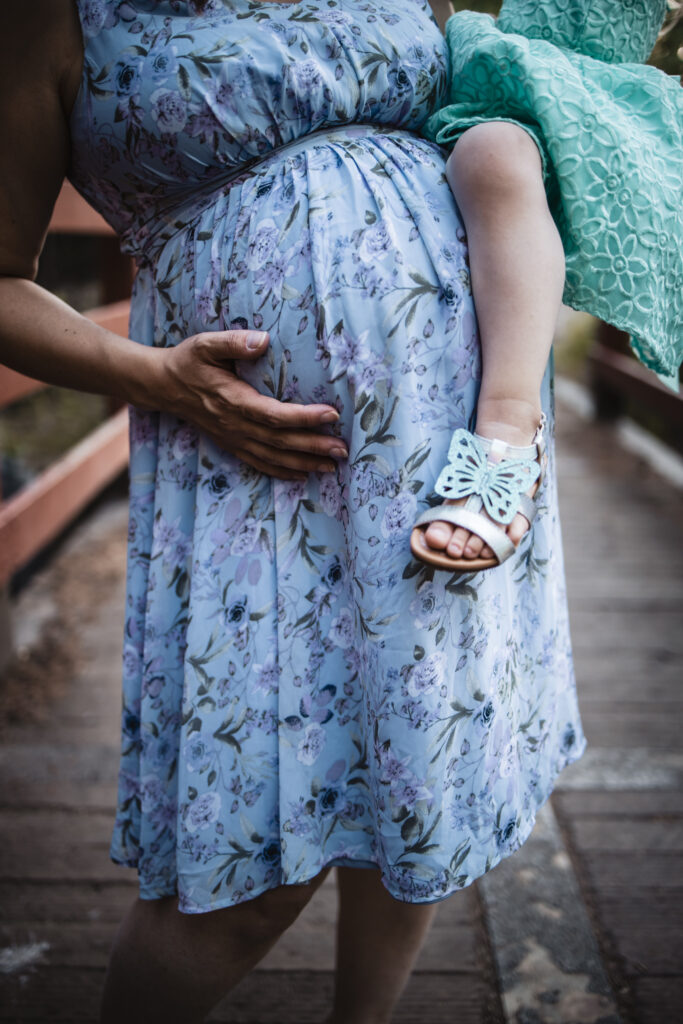 Close-up of a pregnant woman in a blue floral dress holding a child on a rustic bridge.
