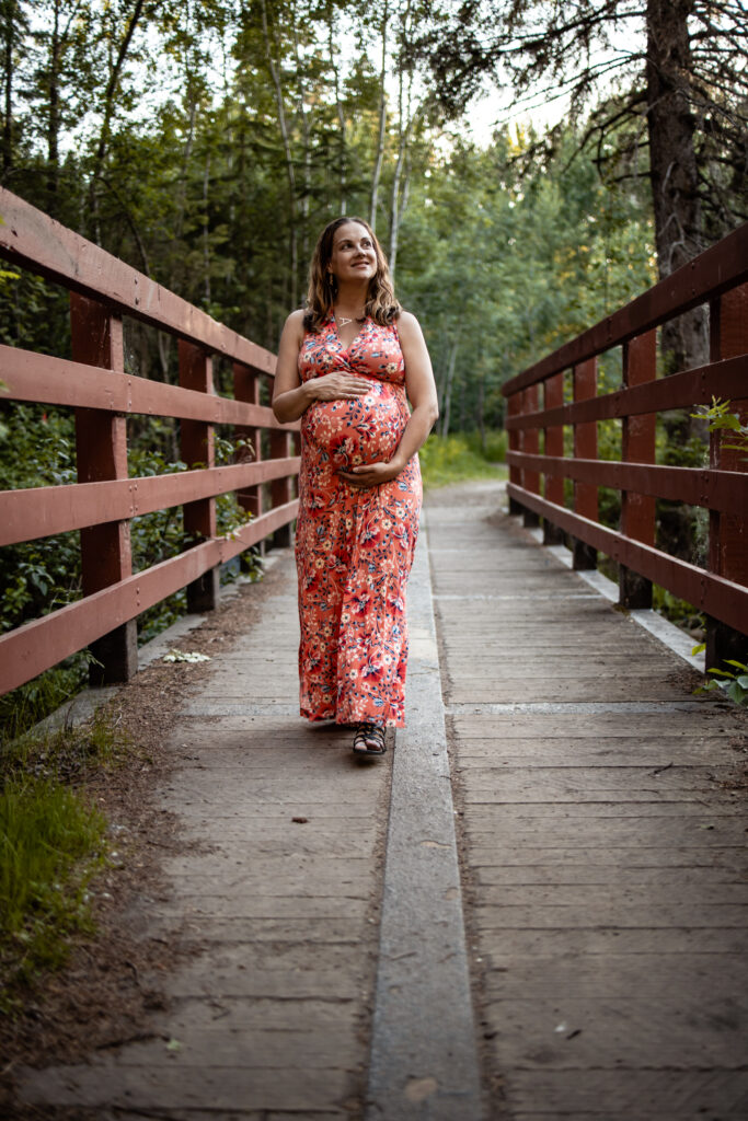 A glowing mama walking across a forest bridge in a coral floral dress — one of the cozy maternity photo outfit ideas for summer sessions.