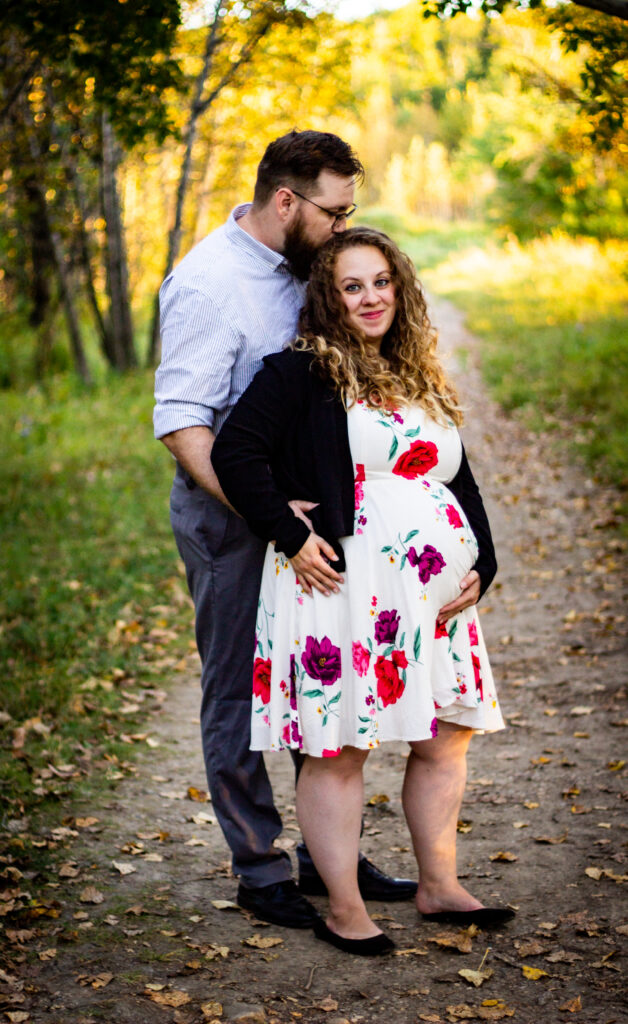 Couple embracing during a maternity shoot in a wooded path, with the mom-to-be in a white floral dress.