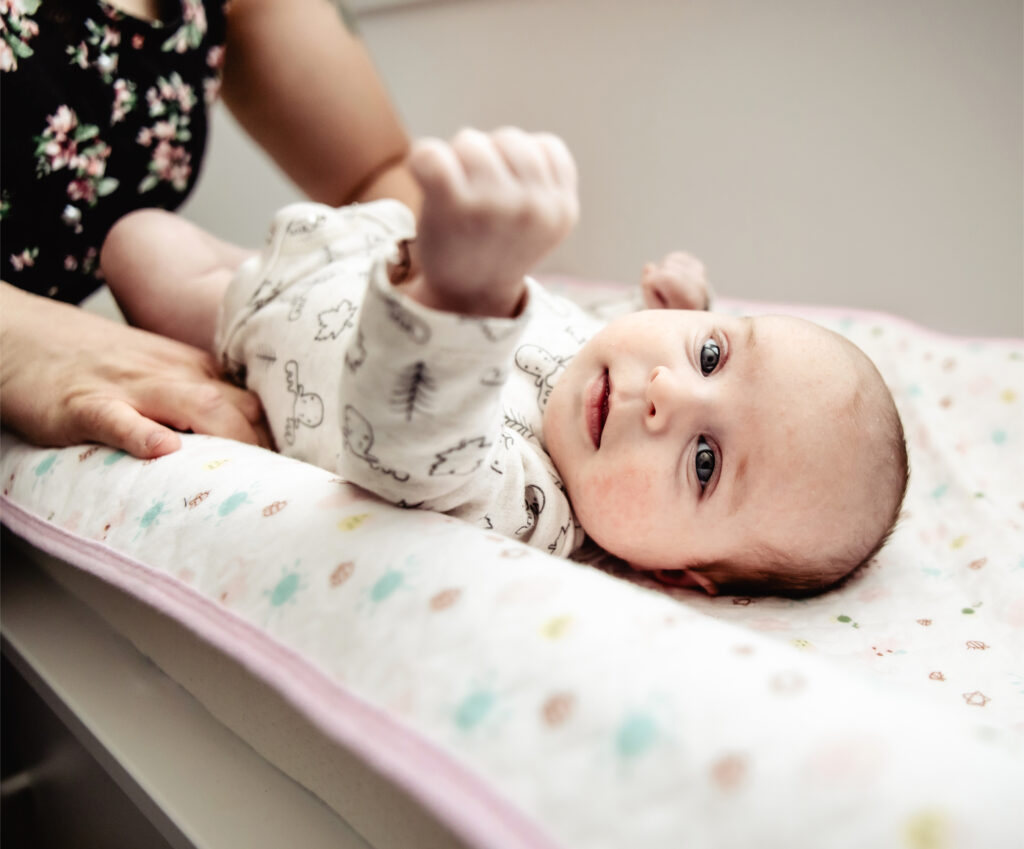A baby lying on a changing pad looking up at the camera while a parent supports them — a candid moment captured in home family photography.