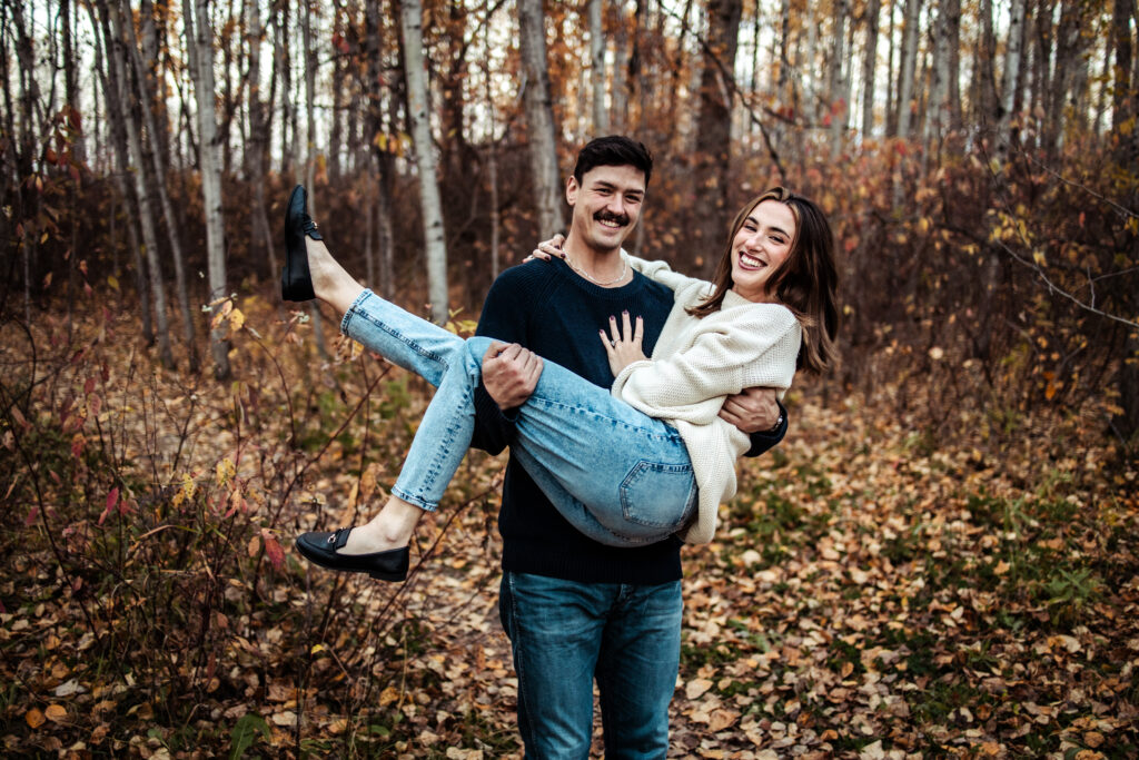 Love story photography: he lifts his laughing partner in an autumn forest, cozy sweaters and jeans, golden leaves underfoot.