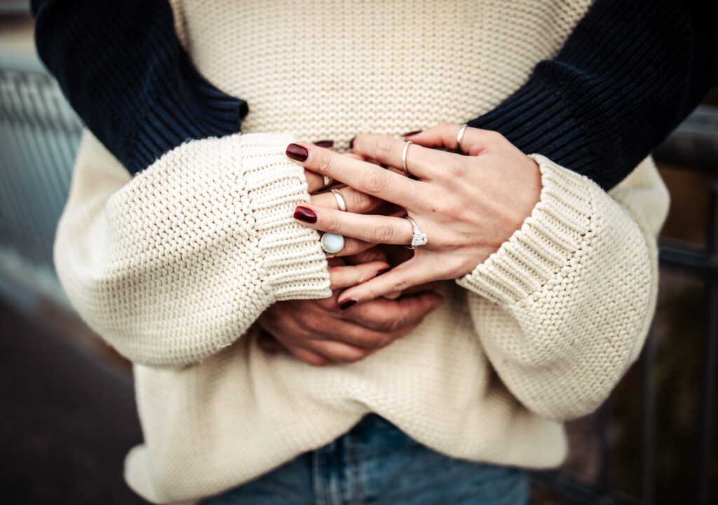 Love story photography: close-up of intertwined hands on a cream sweater, solitaire ring and deep red nails in a warm embrace.