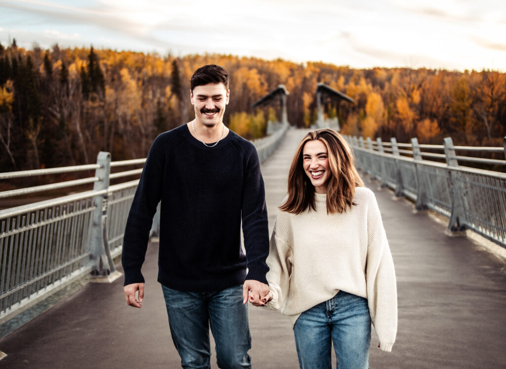 Love story photography: couple walking hand in hand across a bridge at golden hour, big smiles and fall color in the trees.