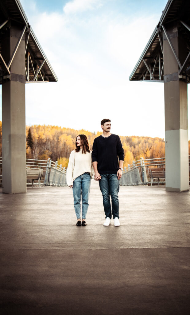 couple holding hands beneath an open-air bridge with wide negative space and golden autumn trees behind.