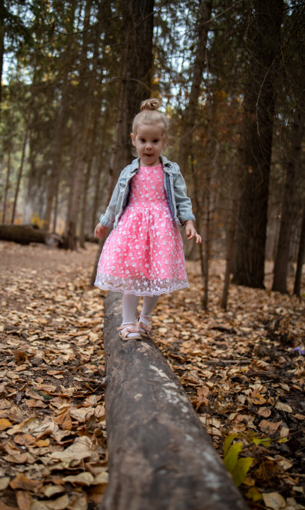 Child balancing on a fallen log in the forest, wearing a pink dress and jacket, surrounded by leaves. A candid autumn moment full of charm and adventure.