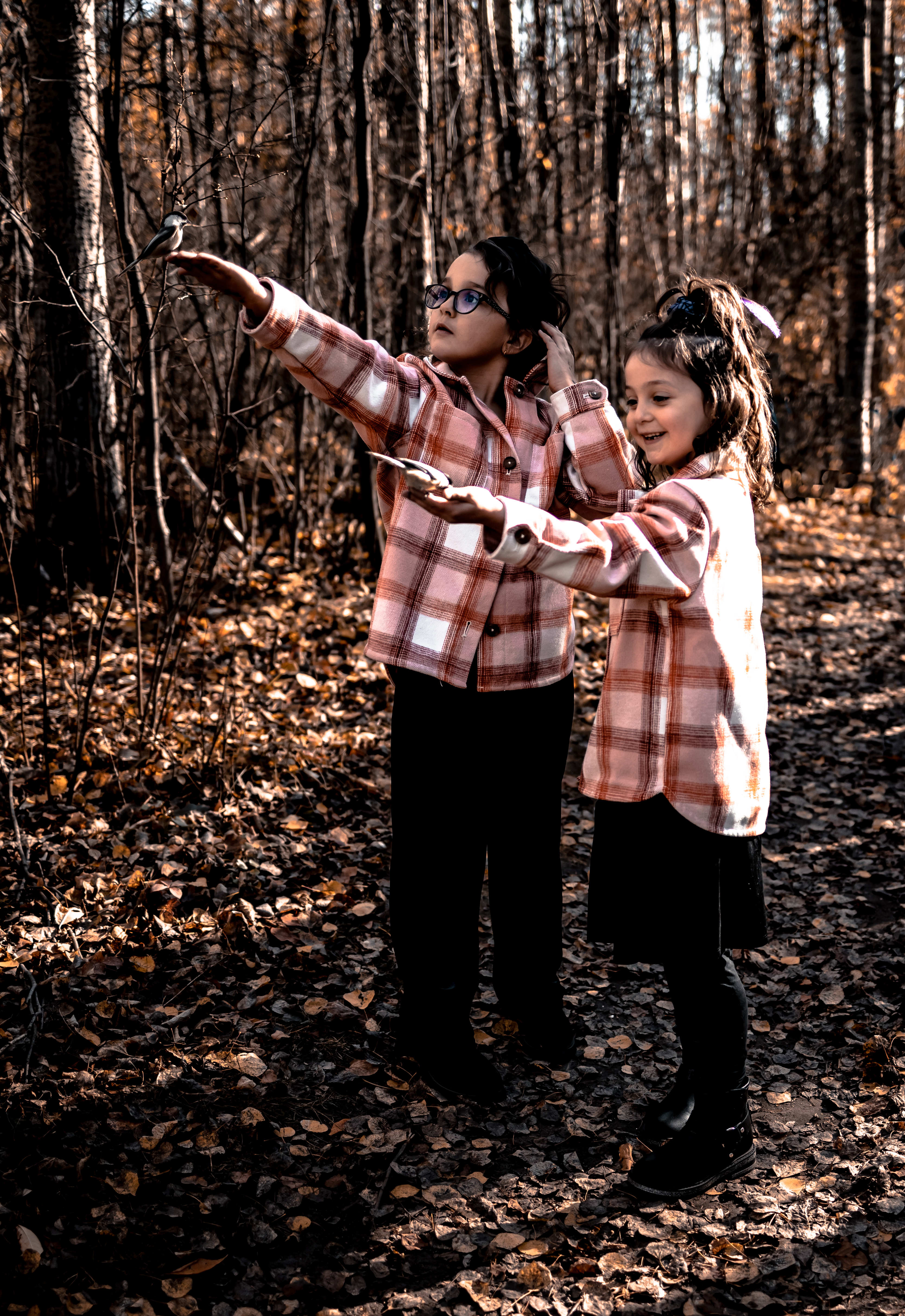 Sisters in matching plaid jackets smiling while feeding birds in the woods, a magical moment for fall family photography ideas.