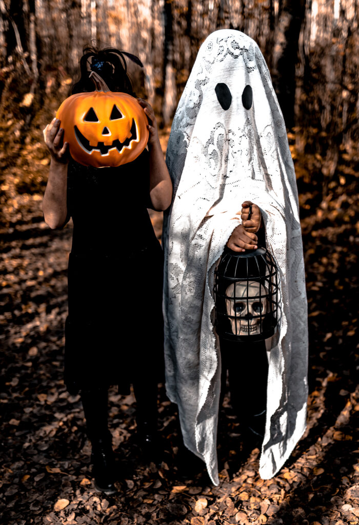 One child dressed as a ghost in a lace sheet holding a caged skull while another hides behind a pumpkin in the woods during autumn. A fun, Halloween-inspired moment.