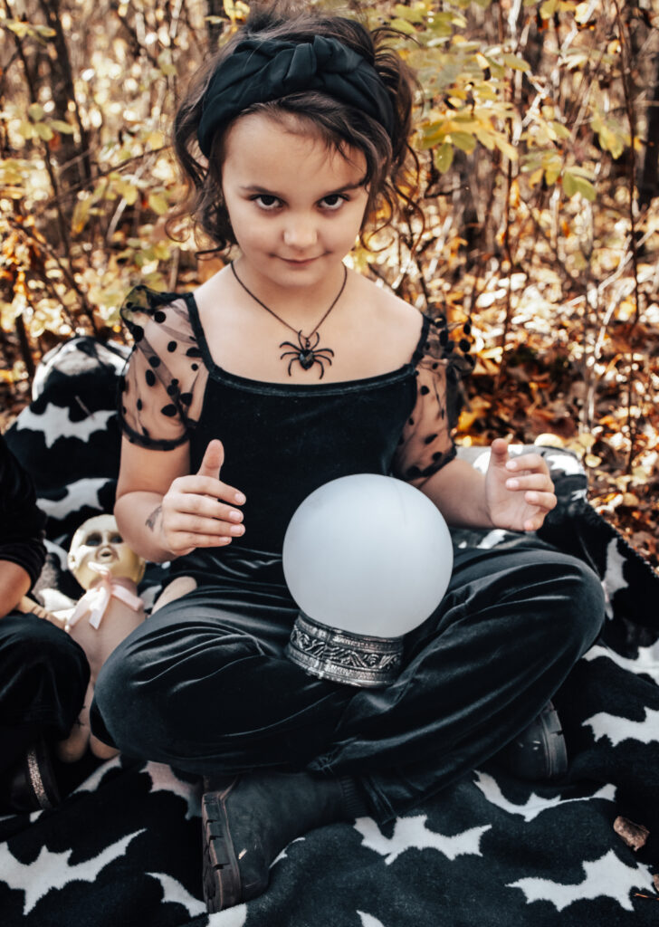 Child in a spider necklace and black outfit sitting cross-legged on a bat blanket, hands hovering over a glowing crystal ball, channeling whimsical fall magic.