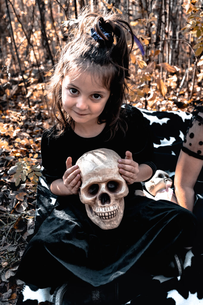 Little girl smiling while holding a skull prop on her lap, surrounded by golden autumn leaves — blending innocence and spooky playfulness for fall family photography ideas.