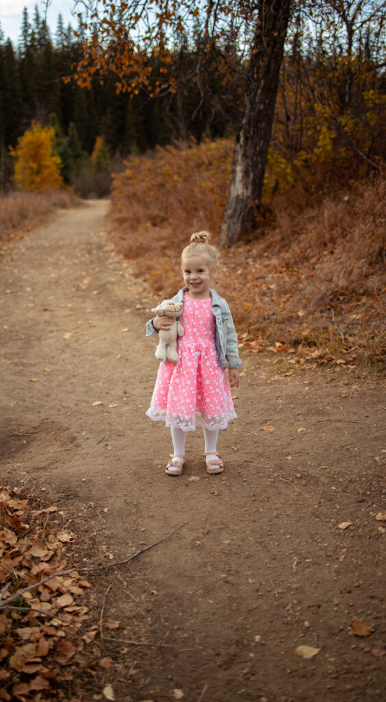 Young girl in a pink dress and denim jacket holding a stuffed animal, standing on a forest path lined with autumn leaves, embodying the sweetness of fall portraits.