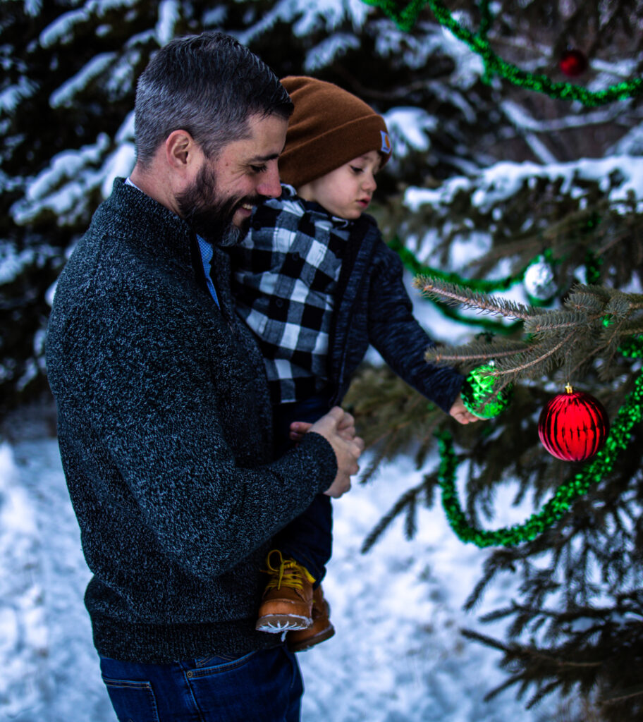 Father holding his young child as they decorate an outdoor Christmas tree during winter Christmas photos Edmonton session.