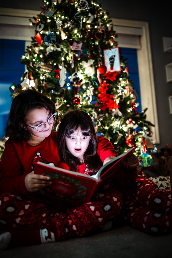 Siblings reading a Christmas story in front of a glowing tree, captured during warm and candid Christmas photos Edmonton session.