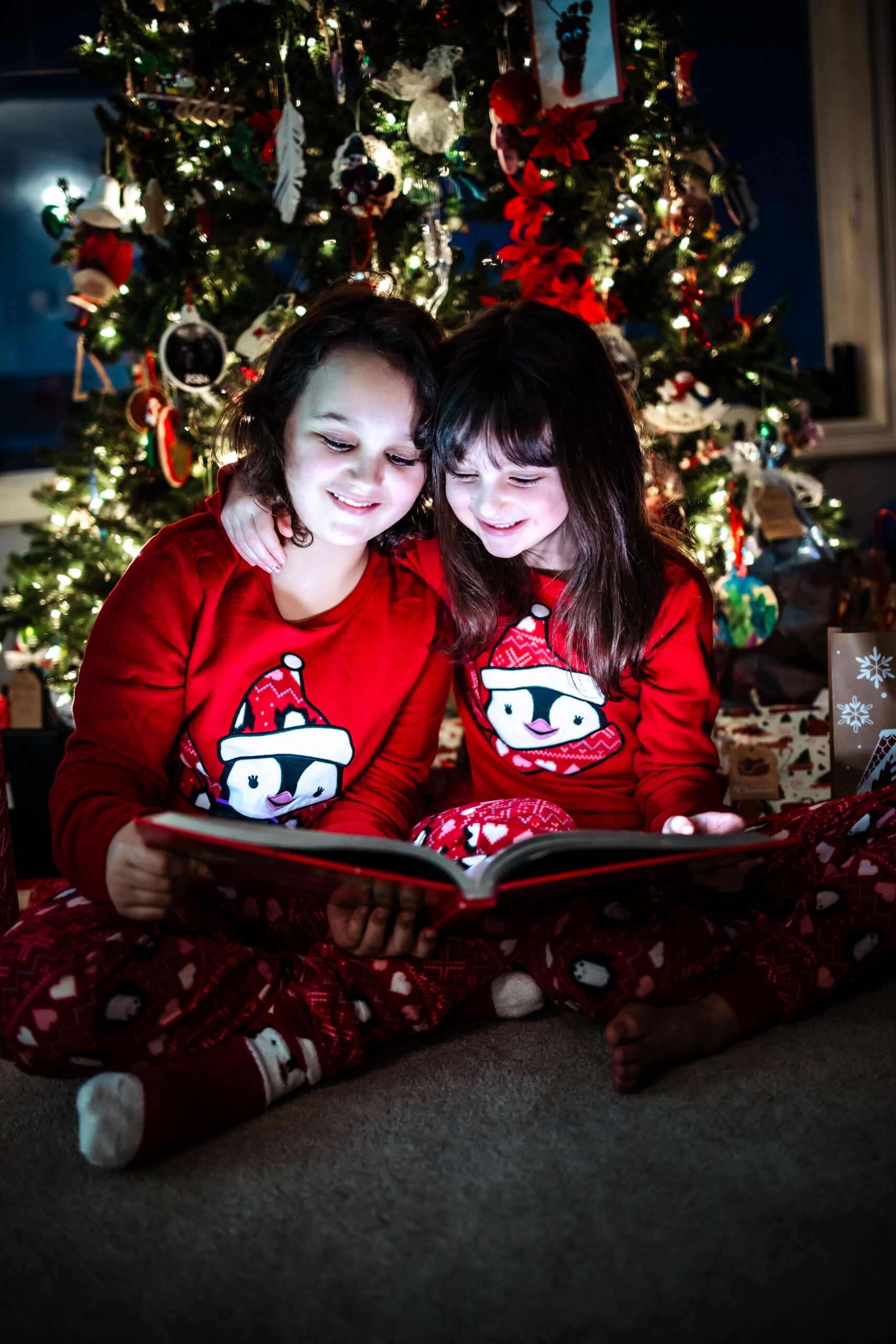 Two children snuggled together reading a holiday book near the Christmas tree for a heartfelt Christmas photos Edmonton session.