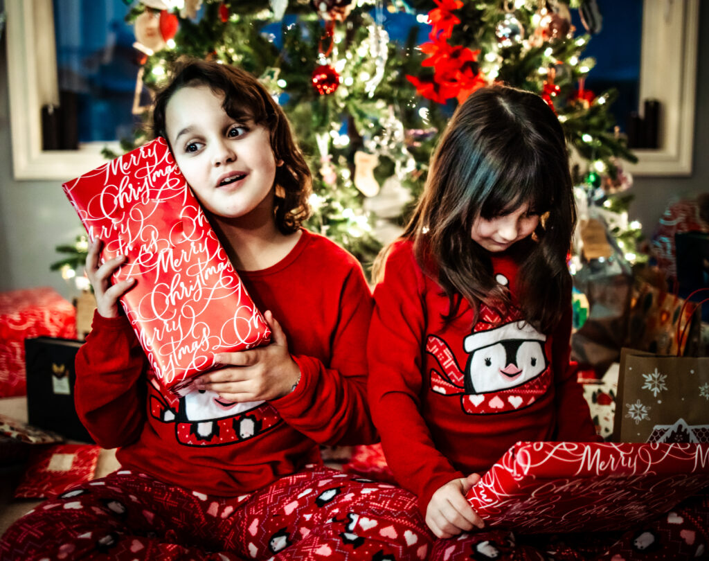 Two children in matching red Christmas pajamas opening presents by the tree during cozy in home photossession.
