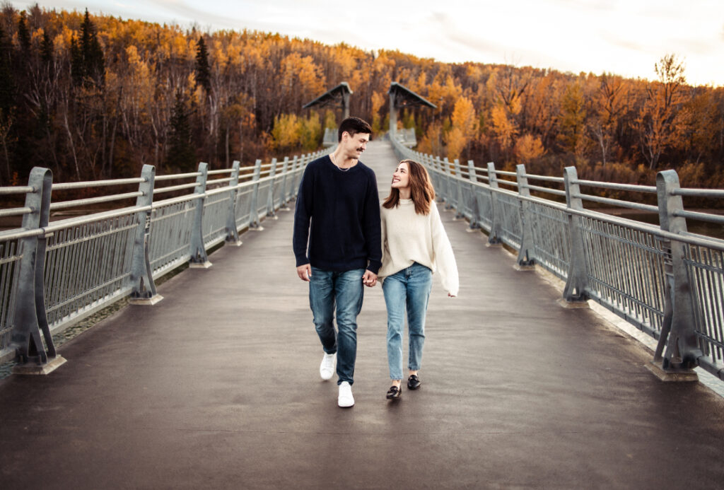 A couple holding hands while walking across a bridge surrounded by autumn trees, photographed at one of the scenic photoshoot locations Edmonton couples love.