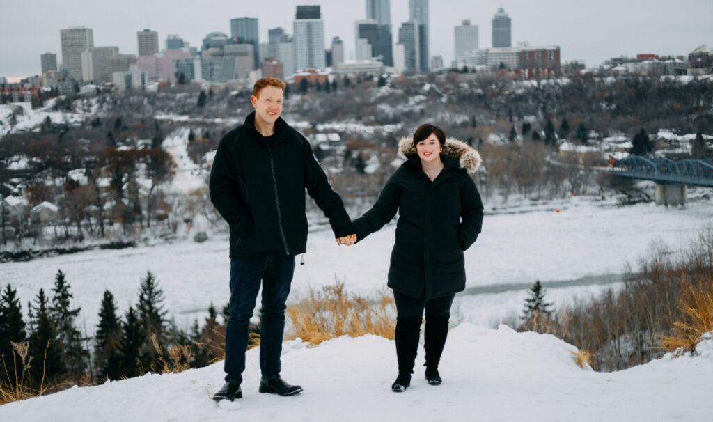 A couple holding hands with the Edmonton skyline behind them, photographed at one of the scenic winter photoshoot locations Edmonton is known for.