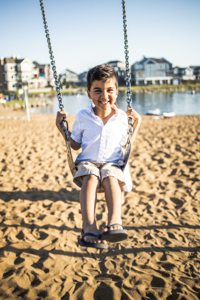 A smiling child sitting on a swing near the water during a summer family session at one of the playful photoshoot locations Edmonton offers.