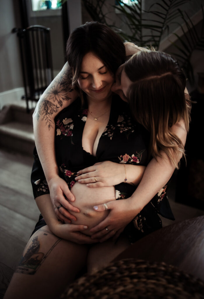 Expectant mother embraces her pregnant belly while her partner hugs her during an intimate maternity session highlighting the support offered by Edmonton doulas and midwives.