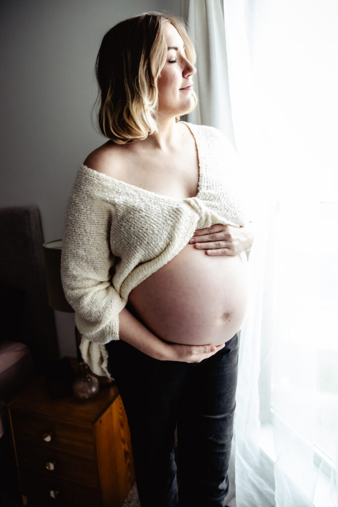 Expectant mother stands beside a bright window, gently cradling her belly during a maternity photography session.