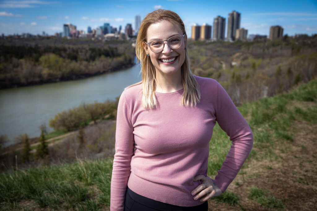 Birth support provider smiles outdoors with the Edmonton skyline behind her, representing the expertise of Edmonton doulas and midwives.