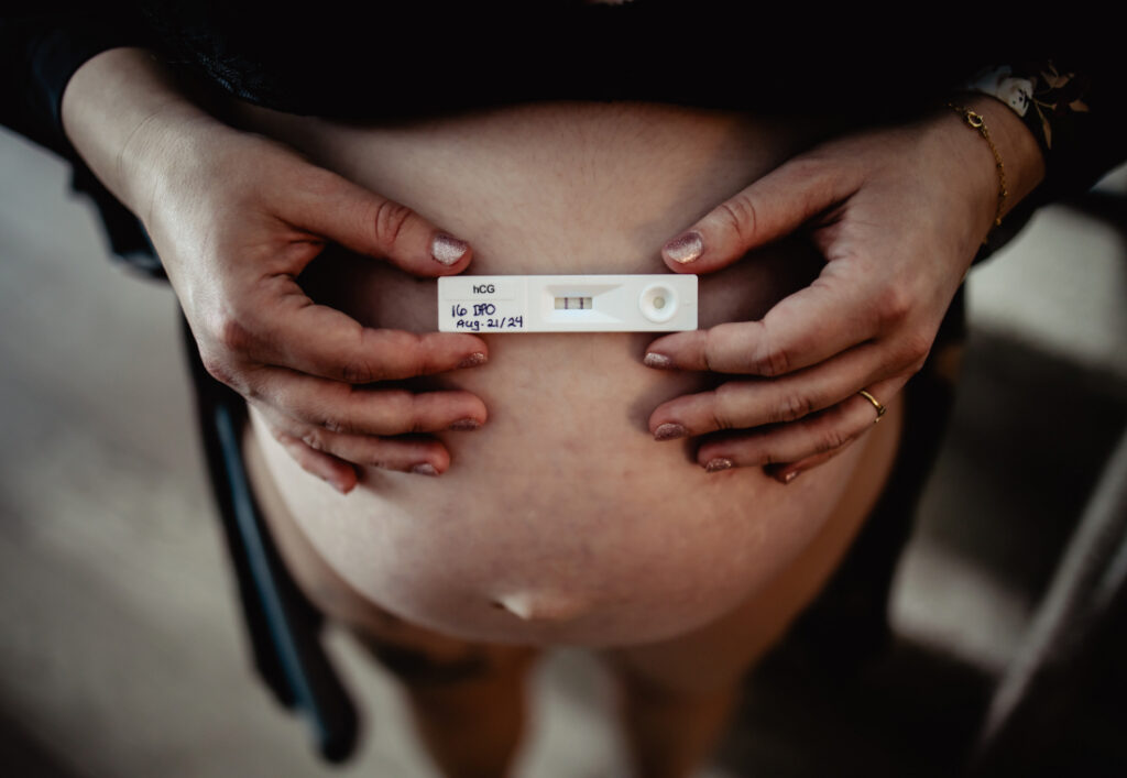 Close-up of a positive pregnancy test resting against a pregnant belly, documenting an early milestone on the journey supported by Edmonton doulas and midwives.