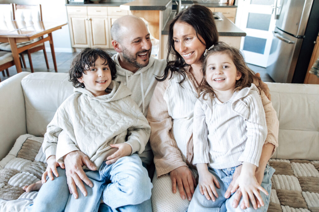 Family of four sits together on a couch, smiling and preparing to welcome a new baby.