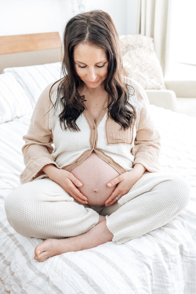 Expectant mother sits cross-legged on a bed, gently holding her belly during a peaceful maternity portrait.